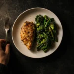 An overhead view of a plate holding a palm-sized portion of chicken and green vegetables, with a hand setting down a fork