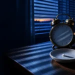 A brass alarm clock and a small ceramic dish on a moonlit bedside table, dawn breaking through the blinds