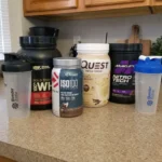 Assorted protein powder tubs and shakers lined up on a kitchen counter